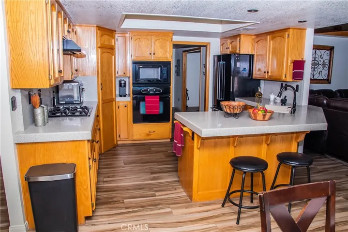 Kitchen showing quartz countertops, large breakfast bar, built in black range/over/dishwasher, cabineted pantry and wood grain tile floors.