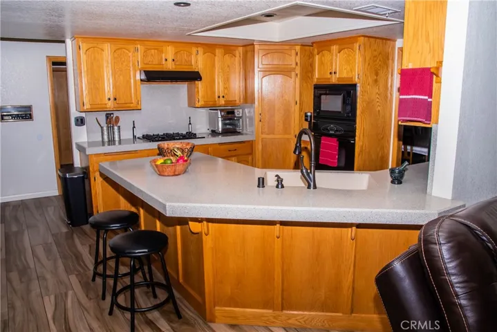 Kitchen showing quartz countertops, large breakfast bar, built in black range/over/dishwasher, cabineted pantry and wood grain tile floors.