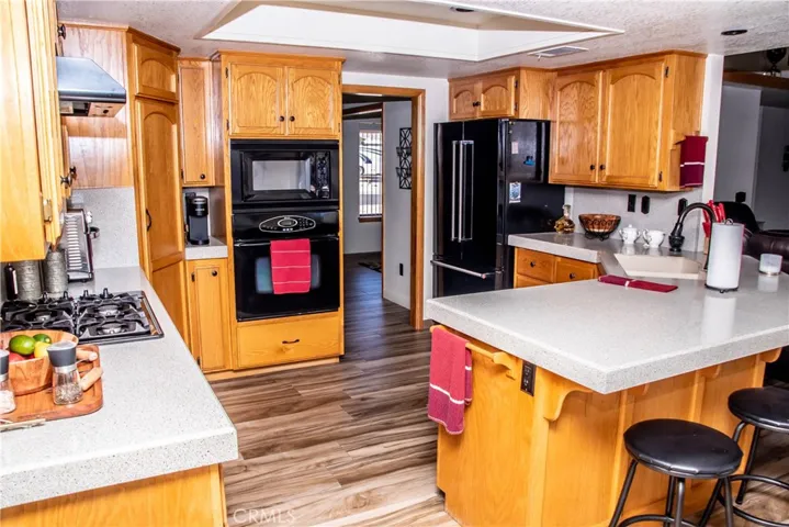 Kitchen showing quartz countertops, large breakfast bar, built in black range/over/dishwasher, cabineted pantry and wood grain tile floors.