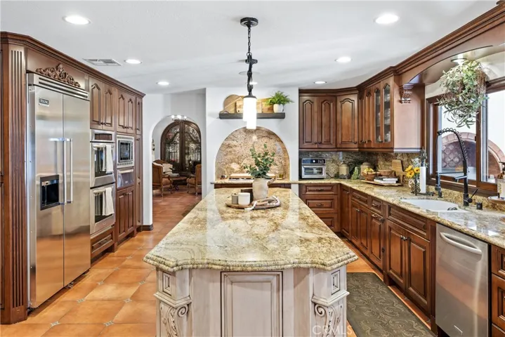 Kitchen featuring custom cabinetry, granite countertops, full granite backsplash, stainless steel Kitchen Aid appliances, center island with pendant lighting, and a sink overlooking the backyard.