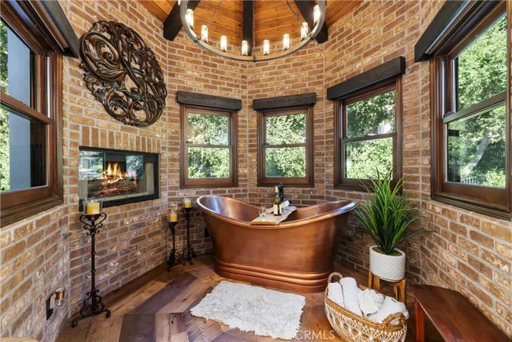 Serene spa alcove featuring a two person free standing copper tub, a fireplace, brick walls, and a beamed ceiling with round chandelier.