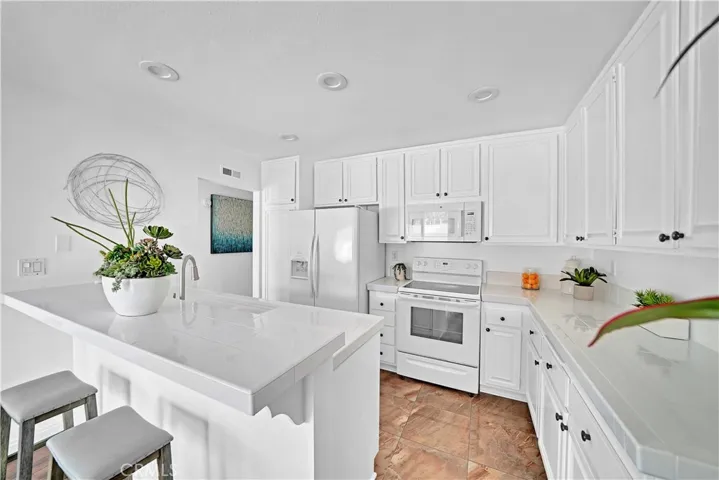 All White Kitchen Boasts Recessed Lighting, white Shaker Cabinetry, Tile Countertops, Breakfast Bar, and White Appliances