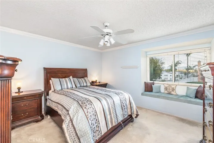 The bedroom offers a tranquil space with a gentle color palette and ample natural light streaming through large windows. A built-in window seat adds a unique architectural detail, while the plush carpeting and a breezy ceiling fan contribute to a comfortable and restful ambiance.