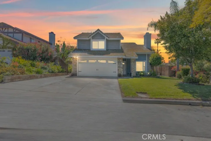 An inviting suburban home with stone accents and a manicured lawn basks in the warm glow of a dusk sky, with a spacious driveway leading to a two-car garage.