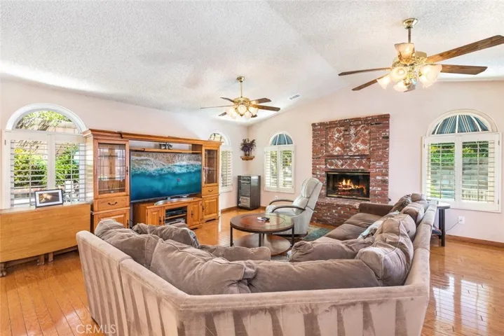 Step into this inviting living room, where the glow of a classic brick fireplace and the gleam of elegant wood floors offer a warm welcome. This room beckons for leisurely days or cozy nights in, while the gentle circulation from ceiling fans and the soft light from shuttered windows create a tranquil, homey haven.