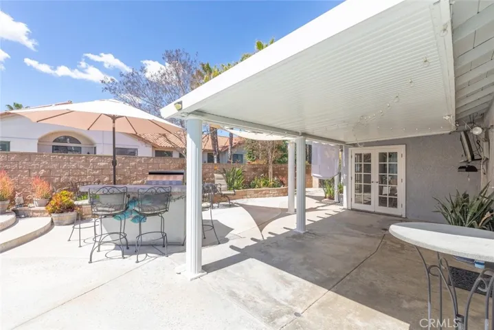 This patio features a large, solid patio cover providing shade over an outdoor dining area.The space is complemented a built-in BBQ grill station against a stone-clad boundary wall. French doors lead into the home, and the overall ambiance is one of a sunny and inviting outdoor living space.
This patio features a large, solid patio cover providing shade over an outdoor dining area with a round table and metal chairs. The space is complemented by potted plants and a built-in BBQ grill station against a sto