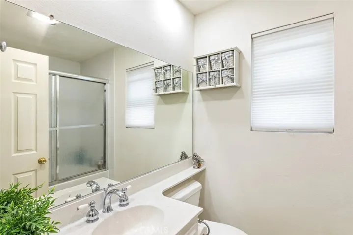 This bathroom is designed with simplicity in mind, featuring a shower-tub combo enclosed by glass doors, a single sink vanity with ample counter space, and a well-lit mirror. Privacy blinds allow for controlled natural light, creating a bright yet private space.