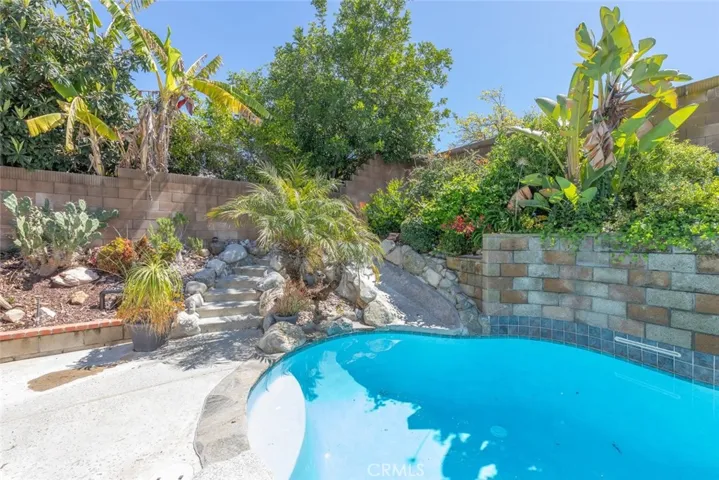 A lush garden landscape with tropical plants and a stone pathway wraps around a curvaceous pool, inviting a leisurely stroll or a refreshing dip.