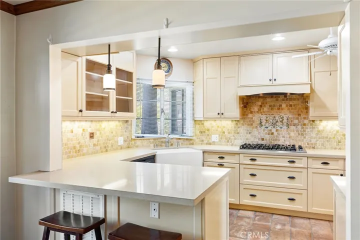 Remodeled kitchen with cream colored cabinetry with inset doors, stone countertops, travertine tiled backsplash, a farm sink, a peninsula with countertop seating and pendant lighting, and a window overlooking the front yard.