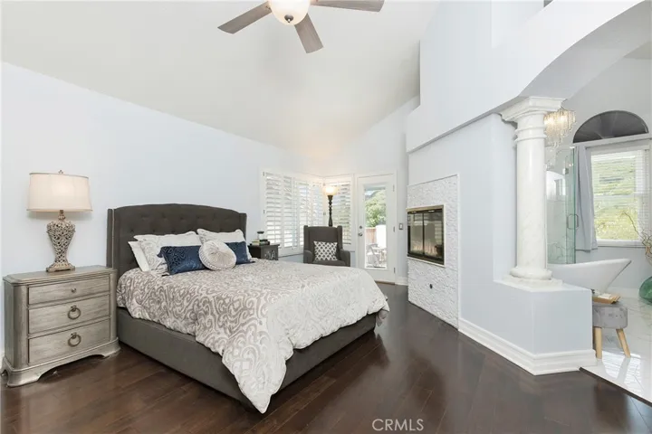 Serene primary bedroom suite with soaring ceilings, ceiling fan, plantation shutters, dark walnut engineered floors, and a romantic 2-sided fireplace.