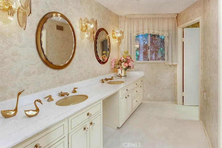Sumptuous en suite primary bathroom with separate dressing area with dual basin vanity with marble countertops, built-in make-up vanity, undermount basins, dual framed dressing mirrors, custom wall sconce lighting, gold-tone fixtures and hardware, and plush wall-to-wall carpet.