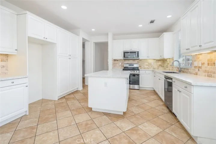 The kitchen features a quartz countertops, a center, white cabinetry, tumbled stone backsplash, and stainless steel appliances.