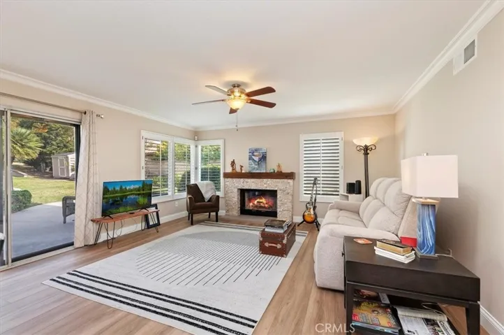 View of the family room. Notice the windows with shutters, ceiling fan with ambience lighting. Sliding patio door to patio area and pool area. Crown moldings and fireplace with stone facia and wooden mantle. Views from windows and slider