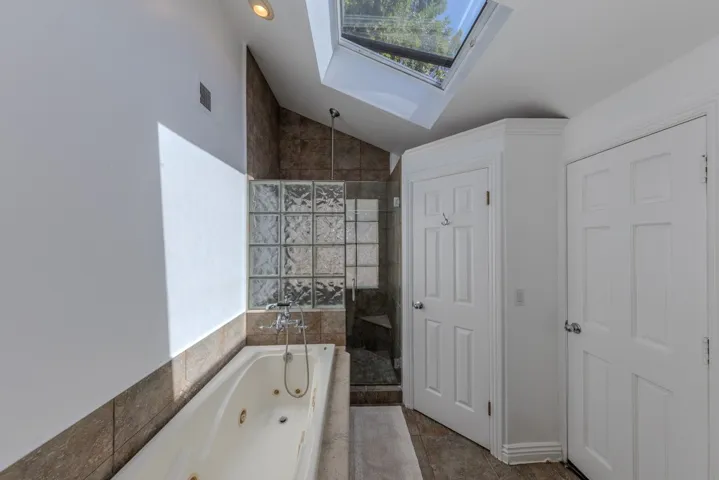 Primary Jacuzzi-brand jetted tub with 2-head stone shower just beyond; second openable giant skylight; note separate toilet room adjacent to shower; note exterior door out to east side yard (which features an outdoor shower)