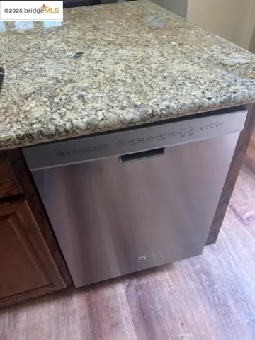 Kitchen view of stainless steel dishwasher, light stone countertops, and wood finished floors