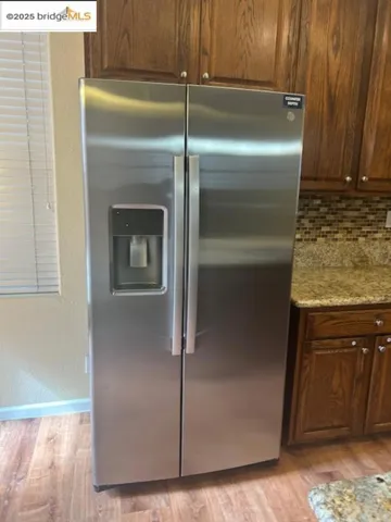 Kitchen with stainless steel fridge, light wood-type flooring, light stone counters, and decorative backsplash