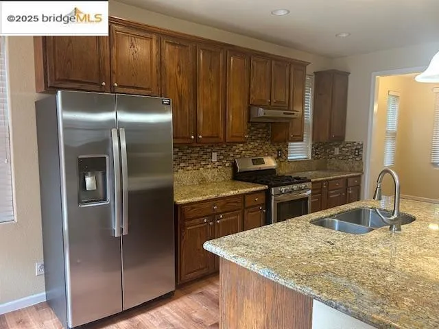 Kitchen featuring stainless steel appliances, light wood finished floors, light stone counters, decorative backsplash, and under cabinet range hood