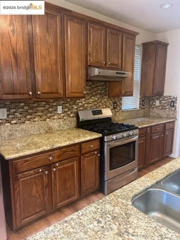 Kitchen with stainless steel gas range, light wood finished floors, under cabinet range hood, decorative backsplash, and light stone countertops