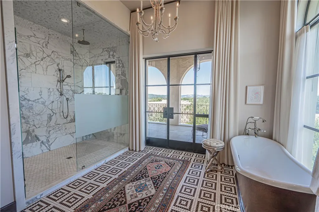 This stunning primary bathroom boasts an expansive marble shower with an adjoining steam shower, a free-standing tub overlooking a picturesque window, a double vanity, and private balcony access through double doors.