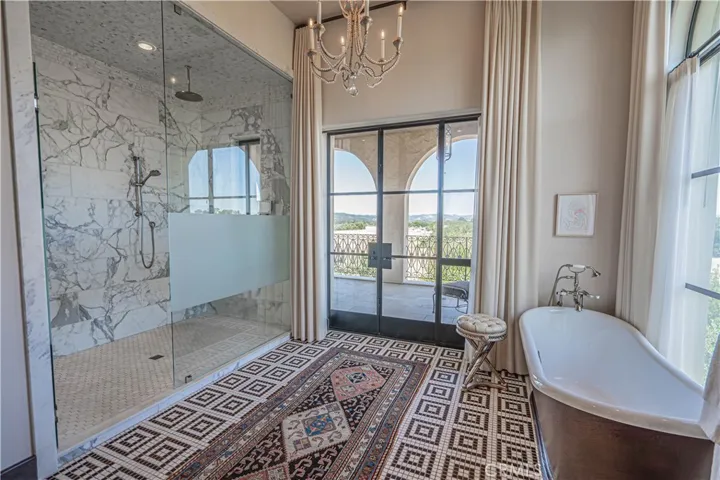 This stunning primary bathroom boasts an expansive marble shower with an adjoining steam shower, a free-standing tub overlooking a picturesque window, a double vanity, and private balcony access through double doors.