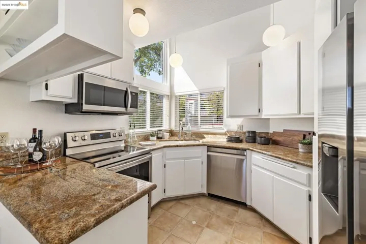 Kitchen with appliances with stainless steel finishes, white cabinets, light tile patterned floors, stone counters, and hanging light fixtures