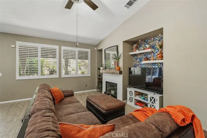 Vaulted ceiling with shutters, ceiling fan, built-in TV niche, and a tiled fireplace featuring a wooden mantle.