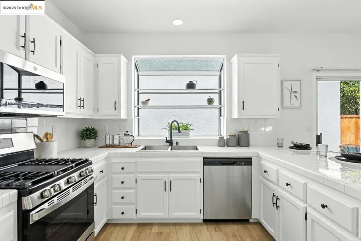 Kitchen with tile counters, appliances with stainless steel finishes, white cabinets, tasteful backsplash, and light wood-style floors