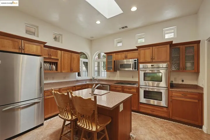 Kitchen featuring stainless steel appliances, brown cabinets, glass insert cabinets, light stone counters, and a skylight