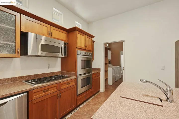 Kitchen featuring appliances with stainless steel finishes, washing machine and clothes dryer, light stone countertops, light tile patterned floors, and brown cabinetry
