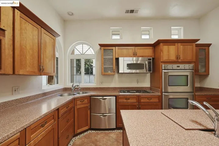 Kitchen with glass insert cabinets, brown cabinetry, stainless steel appliances, light stone counters, and light tile patterned floors
