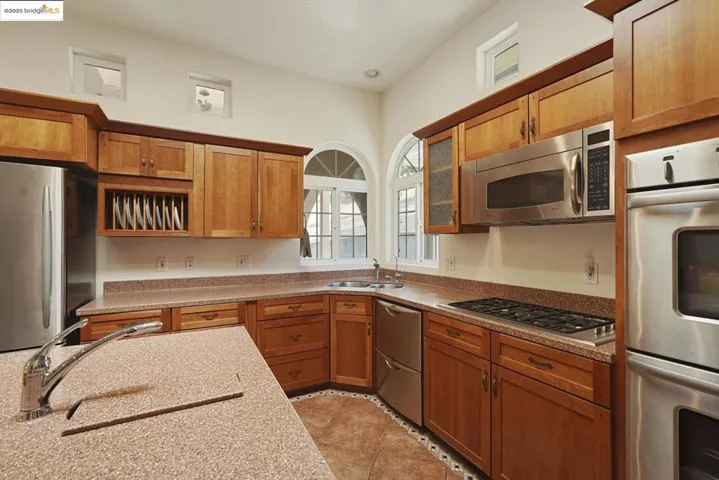 Kitchen with appliances with stainless steel finishes, brown cabinets, light tile patterned flooring, glass insert cabinets, and light stone counters