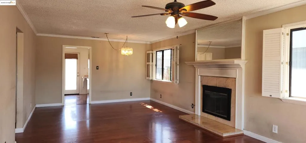 Unfurnished living room with crown molding, a textured ceiling, a tile fireplace, a ceiling fan, and dark wood-style flooring
