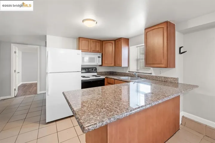 Kitchen with white appliances, light tile patterned floors, light stone counters, a peninsula, and brown cabinetry