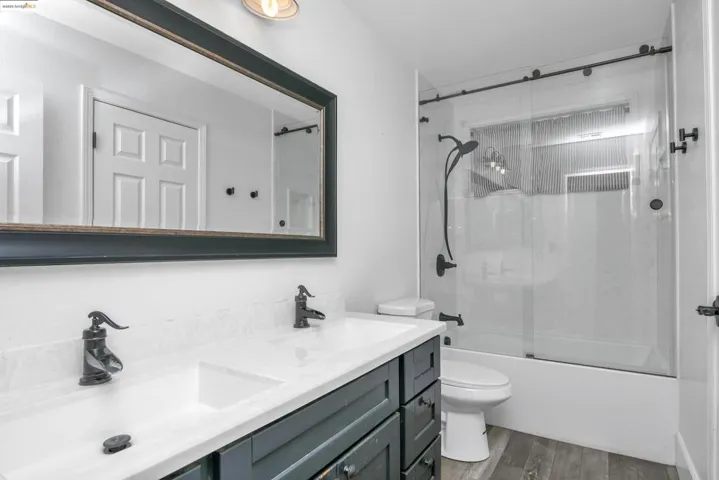 Full bath with light wood finished floors, bath / shower combo with glass door, and double vanity