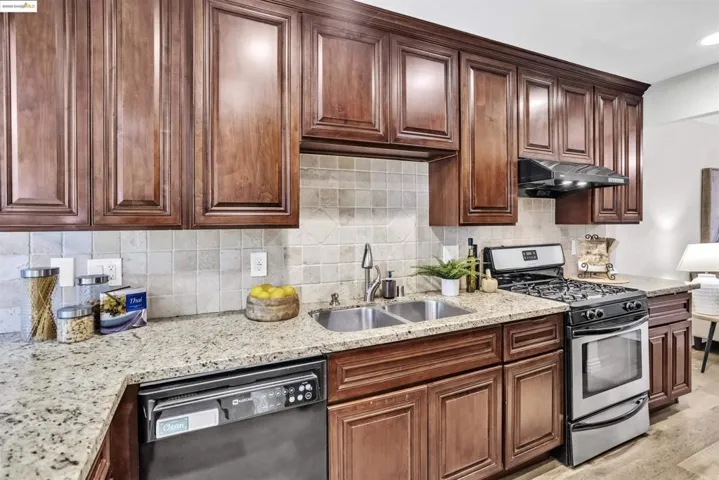 Kitchen with stainless steel gas stove, backsplash, dishwasher, light stone counters, and exhaust hood