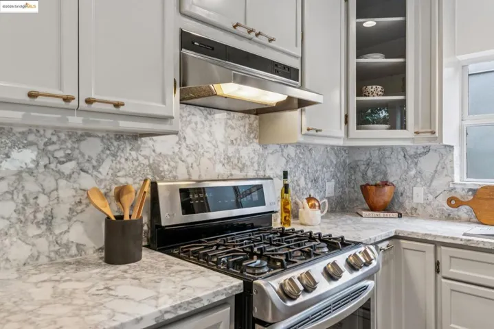 Kitchen with stainless steel gas stove, decorative backsplash, light stone counters, under cabinet range hood, and glass insert cabinets