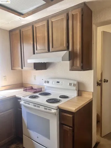 Kitchen featuring a textured wall, white electric stove, light countertops, under cabinet range hood, and dark brown cabinets