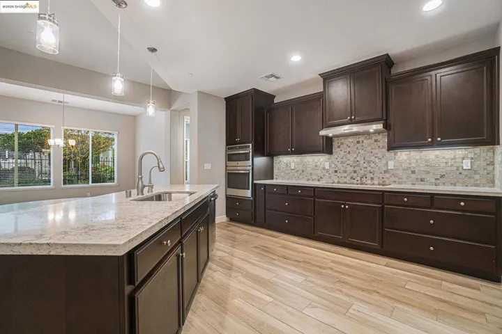 Kitchen featuring dark brown cabinetry, hanging light fixtures, light stone countertops, decorative backsplash, and light wood-style floors