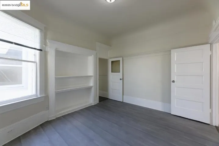 Empty living room with dark wood-type flooring, main window, and doors to the kitchen and hallway to bedrooms and bathroom.