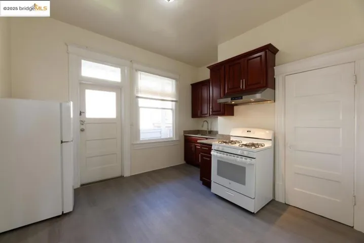 Empty Kitchen with white appliances, dark wood-type flooring, under cabinet range hood, light countertops, and dark cherry wood cabinetry