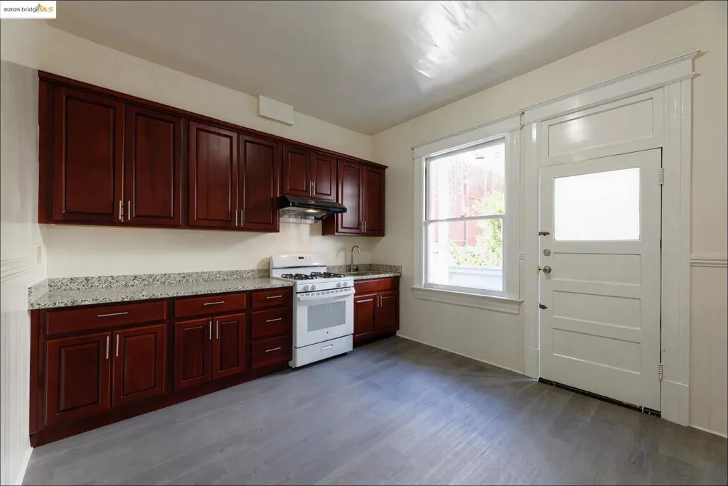 Empty Kitchen featuring white gas range oven, dark wood finished floors, light stone counters, under cabinet range hood, and reddish brown cabinets