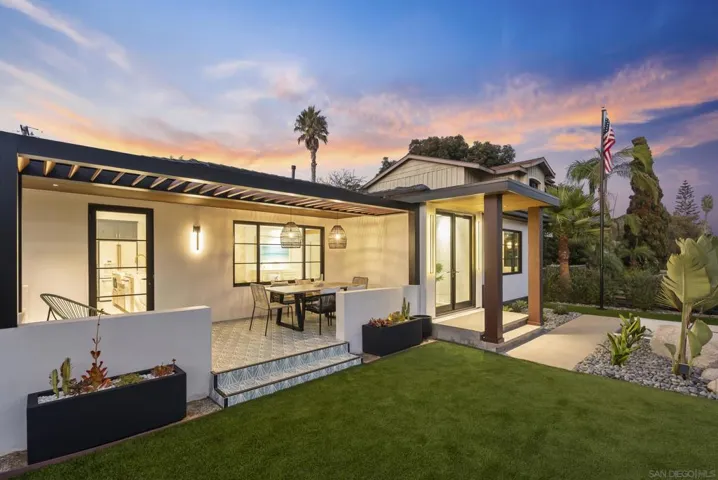 A front-yard dining terrace with a long teak table, subtle privacy hedging, and café lights invites sunset meals in the Pacific breeze.