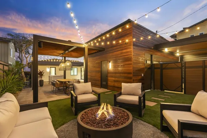 A low stone fire pit with deep lounge seating, flames mirrored in tempered glass, conversation stretching into starry coastal nights.