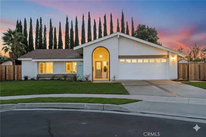 Evening curb appeal with the arched entry aglow, brick-ribbon driveway, fresh landscaping, and cul-de-sac setting under a sunset sky.
