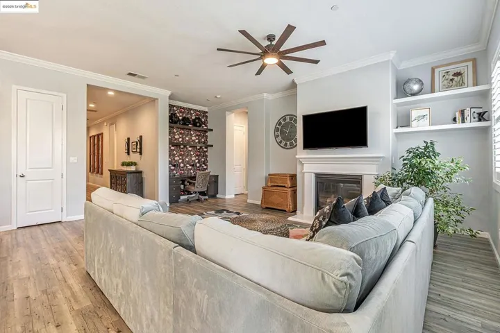 Living room featuring an office area, crown molding, a ceiling fan, a glass covered fireplace, and light wood-style floors