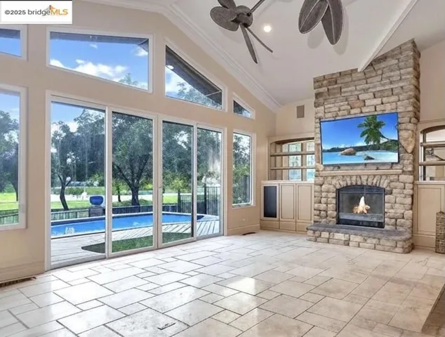 Unfurnished living room with a stone fireplace, ceiling fan, high vaulted ceiling, crown molding, and stone tile floors