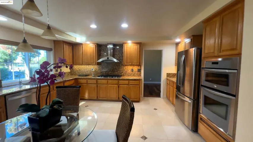 Kitchen with wall chimney range hood, hanging light fixtures, light stone countertops, stainless steel appliances, and brown cabinetry