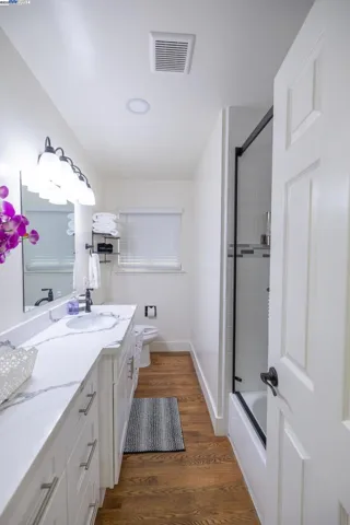 Bright and updated common bathroom featuring a long quartz vanity, modern fixtures, walk-in shower with glass doors, and warm hardwood flooring. Clean, functional, and move-in ready with thoughtful touches throughout.