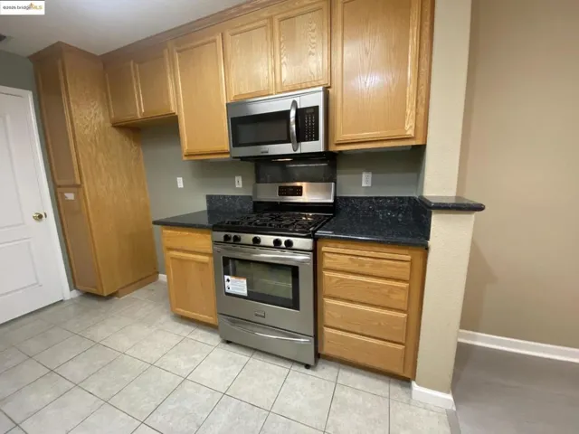 Kitchen featuring stainless steel appliances, dark stone countertops, light tile patterned flooring, and brown cabinetry