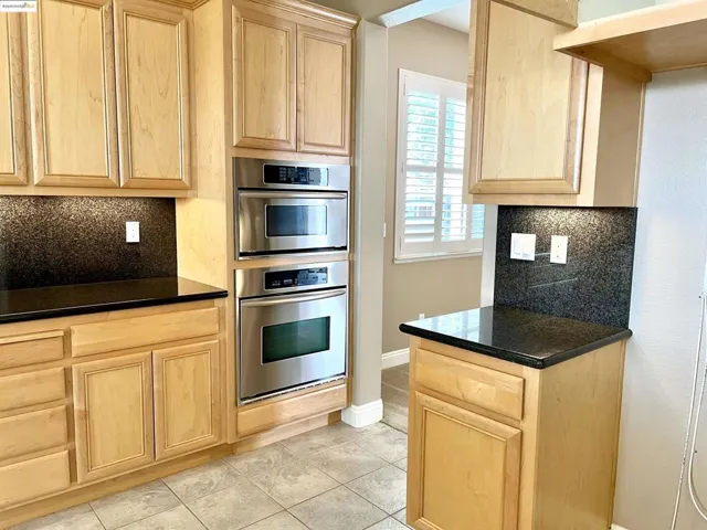 Kitchen with light brown cabinetry, decorative backsplash, double oven, and dark stone countertops